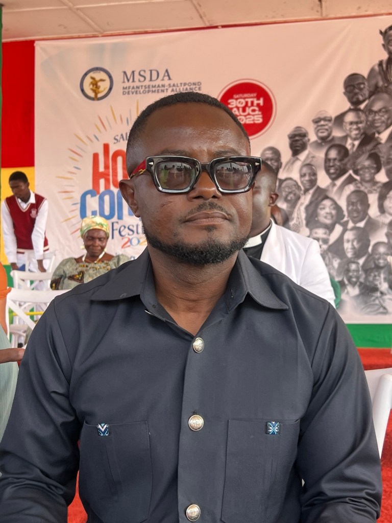 A man wearing sunglasses and a dark shirt is seated, posing for the camera. In the background, there is a colorful banner promoting a community festival with various people appearing in a collage.